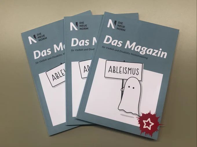 Foto vom Printmagazin Die Neue Norm. Drei Magazine liegen übereinander auf einem Tisch. Auf dem Cover ist ein Geist gezeichnet, der ein Ableismus-Schild hoch hält.