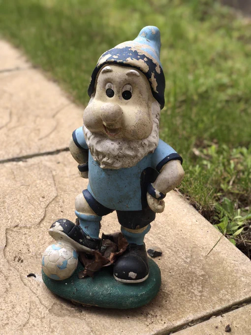 A garden gnome in Coventry City FC football kit with faded and flaking paint, dirty, with leaves around the base.
