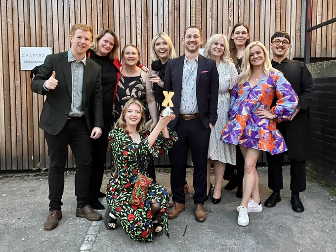 A group of people stand outside and hold up an award in the shape of a silver X.