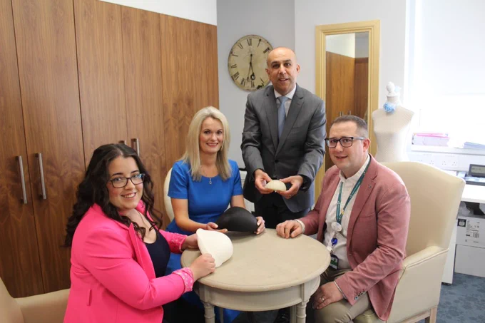 A white room with four people gathered around a small circular table, all wearing smart work clothing. Three are sat down with one standing. They are presenting small breast-shaped prostheses 