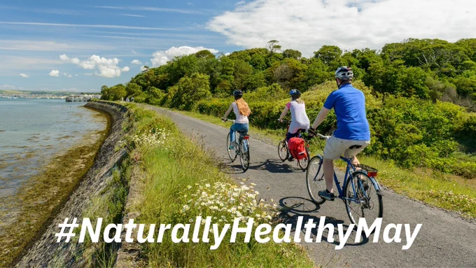 3 people cycling on a coastal path. Text reads: “Naturally Healthy May”