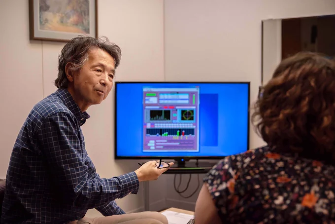 Yoshi speaks to another person sitting next to him in a living room space. A computer monitor in the background shows a screen with multiple charts and settings.