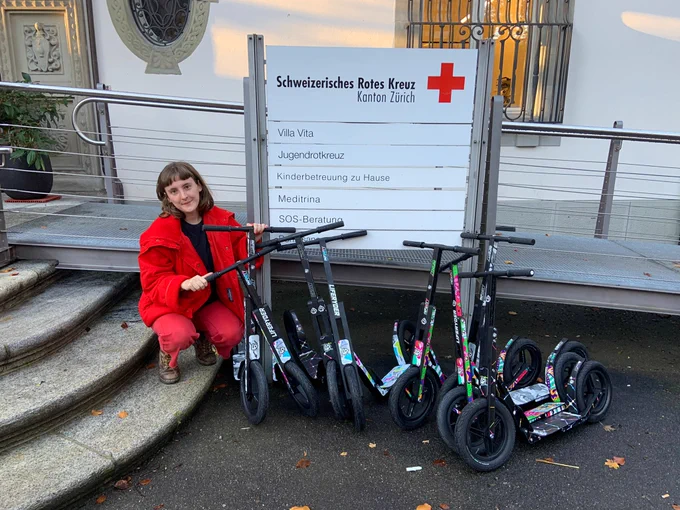 Red Cross Switzerland representative crouches with 7 newly donated scooters