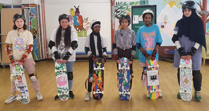 6 young people stand with their new, decorated skateboards whilst wearing helmets, knee, elbow and wrist protection in a community hall