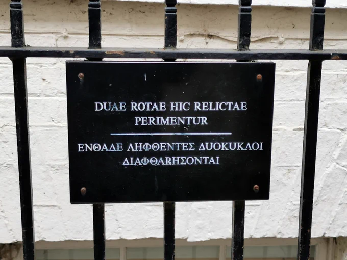 Black sign with white writing in Latin and Greek on black railings: DUAE ROTAE HIC RELICTAE PERIMENTUR - ΕΝΘΑΔΕ ΛΗΦΘΕΝΤΕΣ ΔυΟΚυΚΛΟΙ ΔΙΑΦΘΑΡΗΣΟΝΤΑΙ