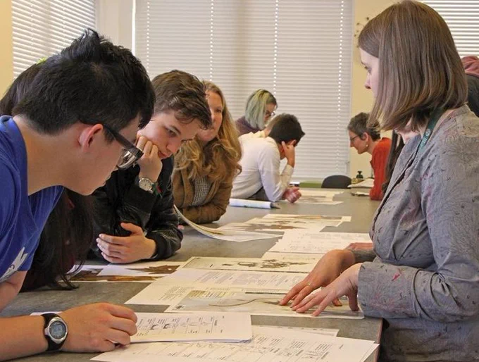 A group of young people sit at a desk looking at plant specimens at a summer camp with a staff member chatting to them.