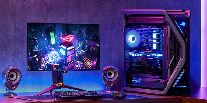 A wide view of a desk setup. There is the ROG PG27AQDM monitor sitting on the desk with two small speakers on either side and a keyboard and mouse in front. To the right of the monitor is the ROG Hyperion Case that's fully built out.