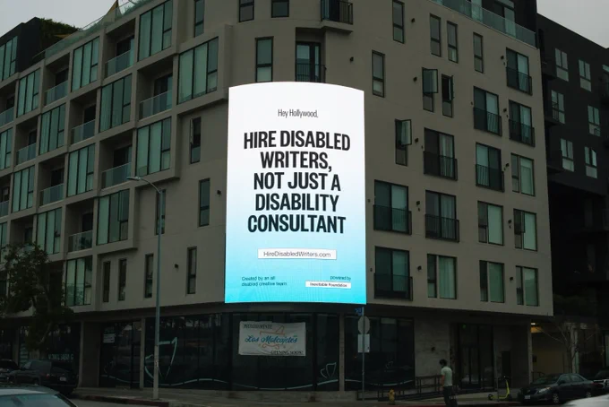 A three story billboard lit up against the side of a building, with bold black text against a blue and white gradient background that reads: Hey Hollywood, Hire Disabled Writers, Not Just A Disability Consultant. Created by an all disabled creative team, and powered by Inevitable Foundation.