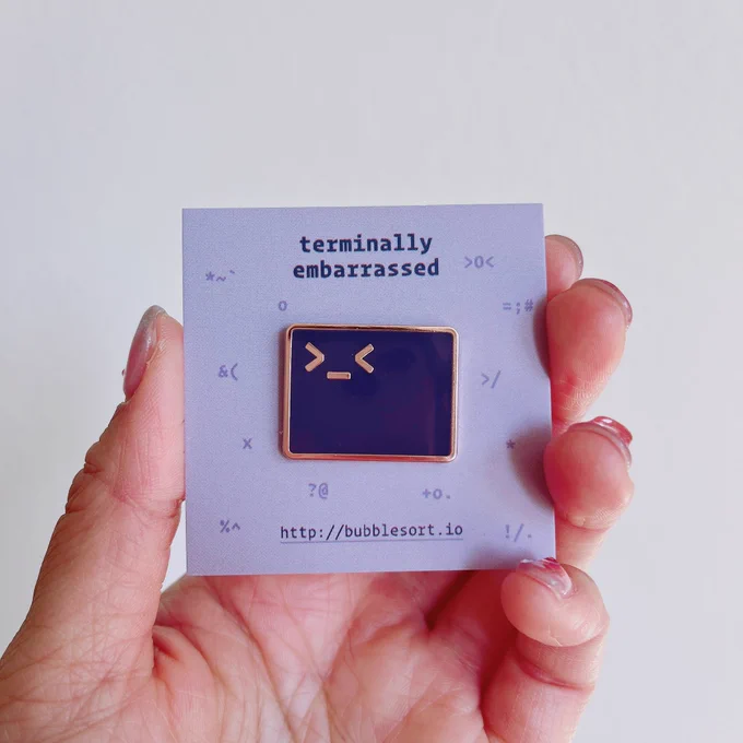 hand holding a backing card with the text “terminally embarrassed” and “bubblesort.io” backing card holds a pin shaped like a terminal, but with an extra < symbol so that the terminal forms an embarrassed wincing face. the terminal is dark purple and the pin has rose gold edging.