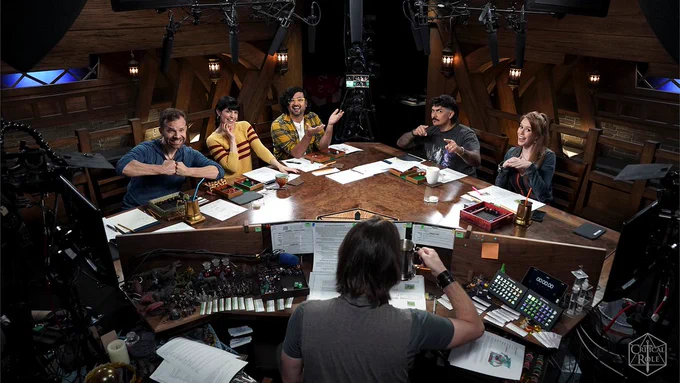 A photo of the cast around the wooden CR table. From left to right it features Liam O'Brien, Emily Axford, Omar Najam, Robbie Daymond, and Marisha Ray. Matthew Mercer sits behind the GM screen with several papers and dice. 