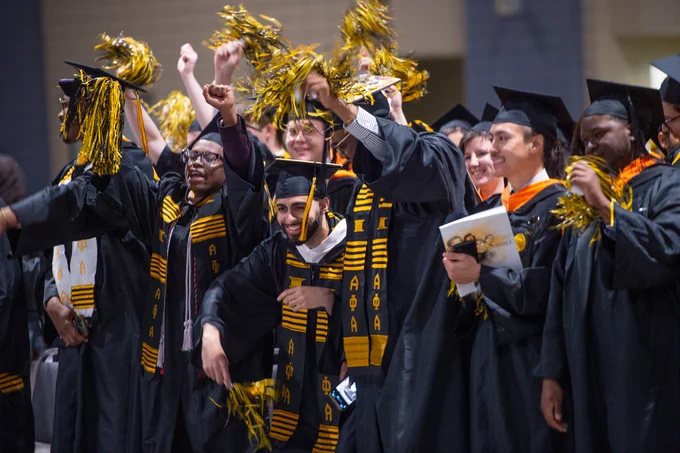 Students in caps and gowns wave yellow and black pom poms.