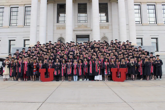 A large group of Master of Social Work students wearing graduation regalia hold their hands in a "U" shape while standing on the steps of the Park Building, surrounding two large, red block U's.