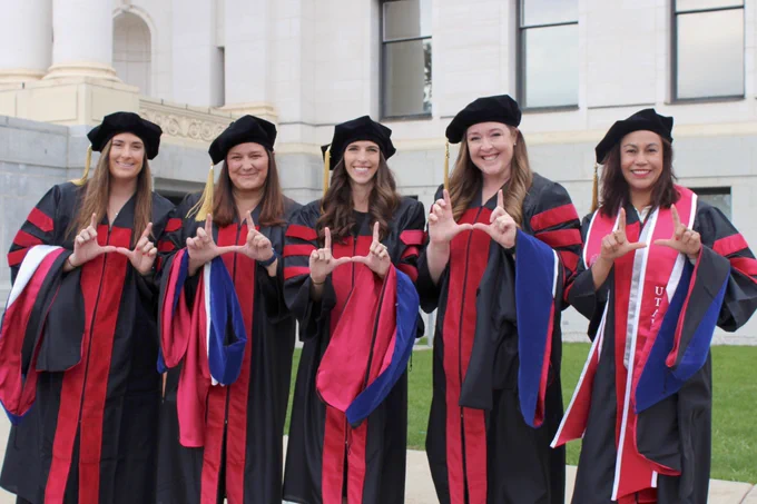 Five smiling social work PhD students wearing graduation regalia, with their doctoral hoods draped over their arms, stand in front of the Park Building and make a "U" shape with their hands.