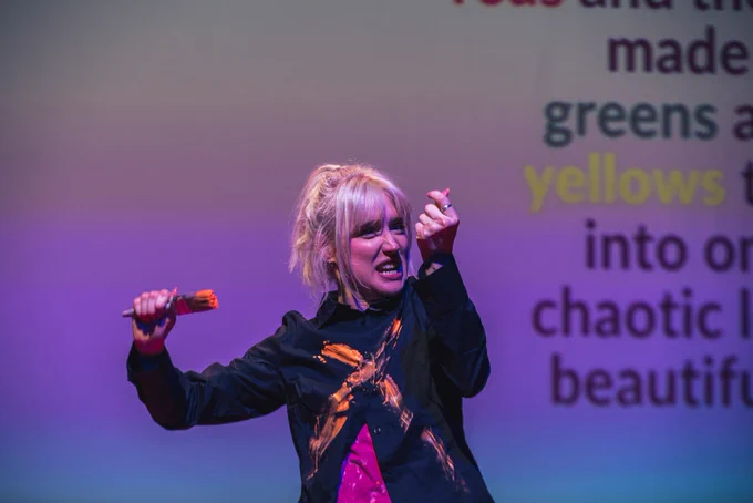 PRODUCTION SHOT. Alice in a black shitt w a painted orange c stands w a orange paint brush in one hand whilst focusing her gaze on her right hand in a fist