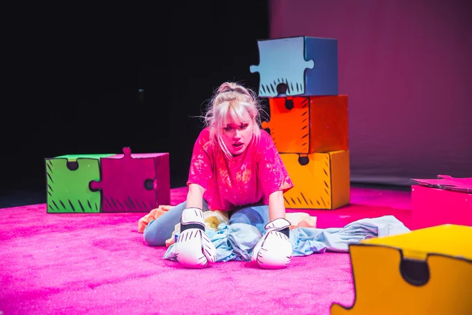 PRODUCTION SHOT. Alice knelt on the pink floor w white boxing gloves stares at the floor surrounded by blue and orange bed sheets and colourful jigsaw squares
