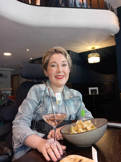 Sarah is sat at a table in a restaurant smiling at the camera. She is holding a glass of rose wine with a straw and has a bowl of salad and tofu. The decor is dark teal, dark wood and gold. Sarah is white, with short blonde hair, wearing a denim jacket and red lipstick