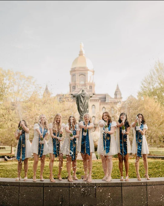 Eight senior women pop champagne in front of the Golden Dome