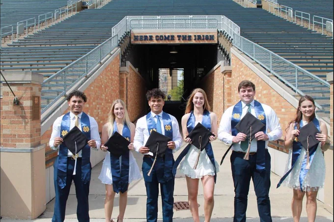 Six seniors stand in front of "Here Come The Irish" tunnel in Notre Dame Stadium while wearing their stoles and holding their graduation caps.