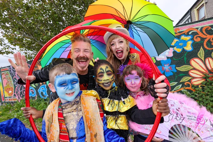 A group of 2 adults and 3 children stand posing with rainbow umbrellas, behind a red hula hoop. The children have their faces painted in bright colours. 