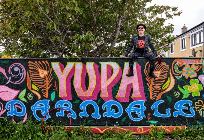 A person sits atop a wall with bright graffiti with designs and graphic text reading: YUPA DARNDALE