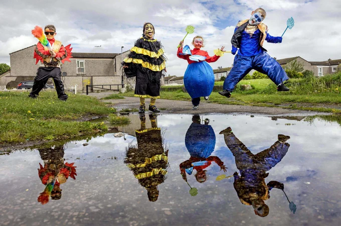 4 young people in different bright costumes post and jump outdoors in a field. Their reflections are seen in a puddle on the ground. 