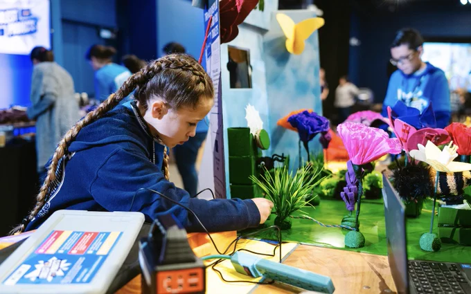 A young female creator surrounded by tech and flowers at an in-person Coolest Projects event.