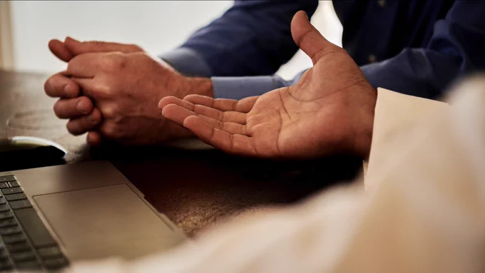A pair of hands is clasped on a table next to a laptop. Another person's open palm gestures towards them.