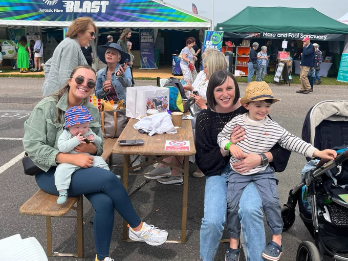 Two adults, each holding a child, sat around a picnic table, facing the camera. Others are sitting in the background.