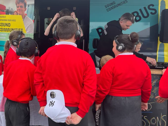 Group of school children with their backs to the camera, with headphones on, gathered around the Sound Communities stand listening to music.