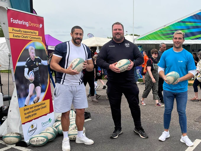 3 adults holding rugby balls, standing in front of a sign saying, Fostering Devon.