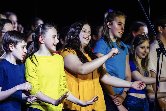 A group of young people wearing brightly coloured tshirts and jeans wave their arms in front of them while singing.