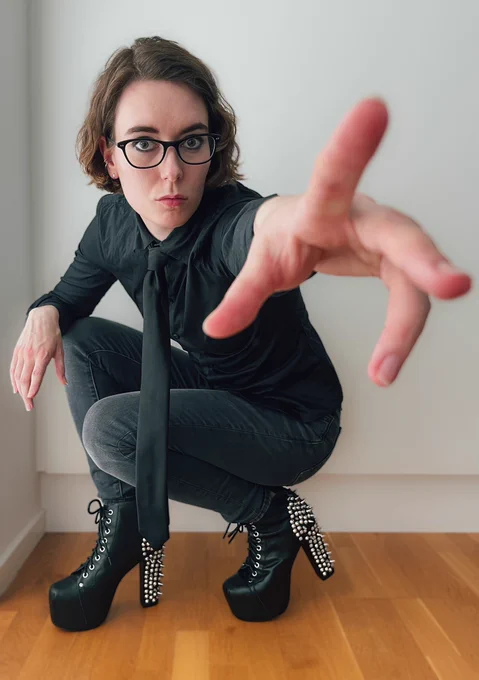 Pind in a black shirt and black tie, gray jeans and studded black leather shoes. She’s crouched down and reaching for the future the patriarchy doesn’t want her to get.