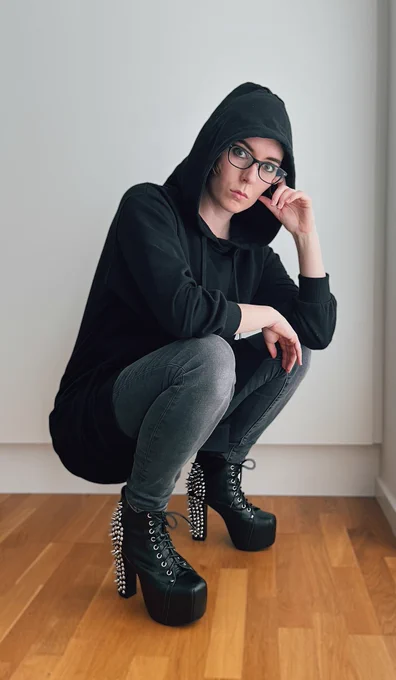 Pind in a long black hoodie, gray jeans and studded black leather heels crouching down with her head resting on one hand.