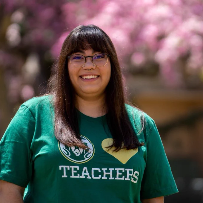 Litzy wears a 'Cam loves teachers' t-shirt in front of a blooming tree with pink flowers. 