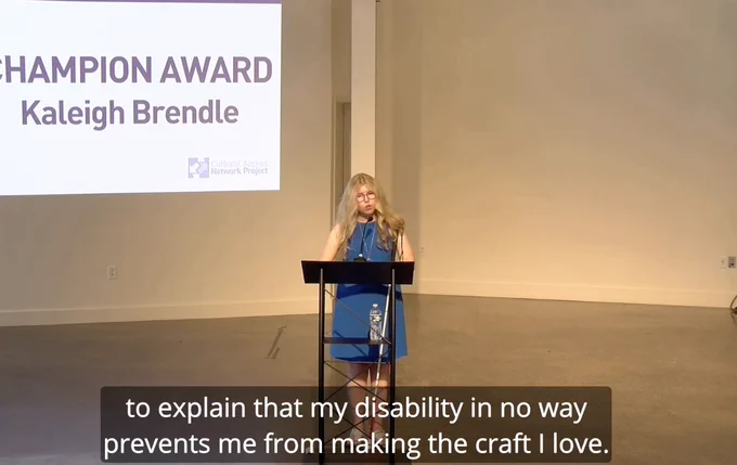 Keleigh, a young white blonde female speaks at an award ceremony with cc text reading: my disability in no way prevents me from making the craft I love