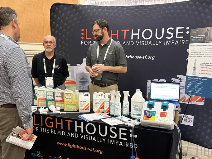 LightHuse COO Brandon Cox and Senior Director of Products, Al Ahlm, stand at the LightHouse booth with a branded backdrop and table lined with LightHouse Industry products are on display