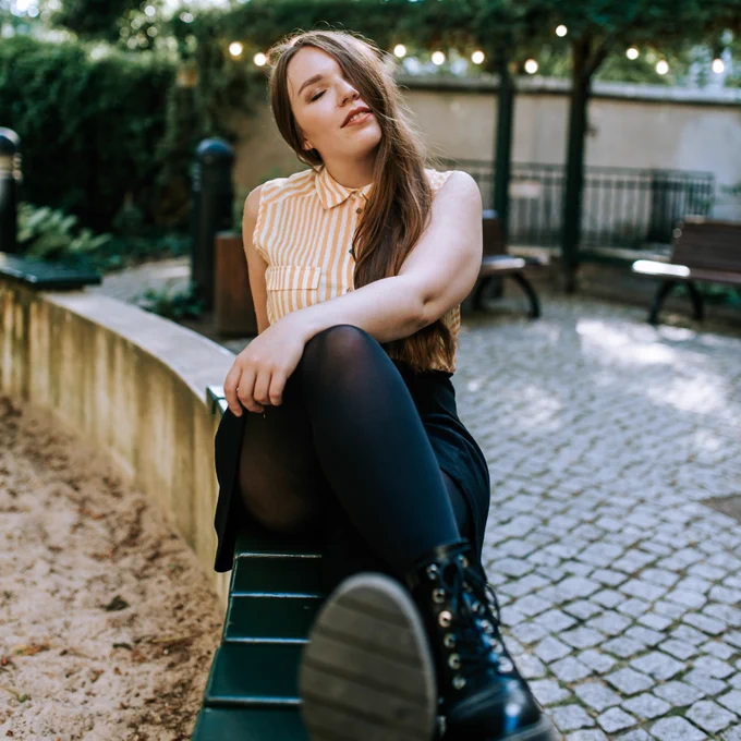 Ich trage einen schwarzen Rock, eine schwarze Strumpfhose, schwarze Schuhe und eine gelb-weiße Bluse. Ich sitze in einem Innenhof auf einer kleinen Mauer und mein Körper ist direkt zur Kamera gedreht. Im Hintergrund sieht man in der Unschärfe ein paar Bänke und eine Lichterkette, die in Bäumen hängt. Meine Haare bedecken die Hälfte meines Gesichts und mein Blick schweift mit geschlossenen Augen leicht nach oben. Meine Hand lege ich auf meinem Knie ab. 

English: I am wearing a black skirt, black tights, black shoes and a yellow and white blouse. I am sitting in a courtyard on a small wall and my body is turned directly towards the camera. In the background, you can see a few benches and a string of lights hanging in trees in the blur. My hair covers half of my face and my gaze wanders slightly upward with my eyes closed. I rest my hand on my knee. 