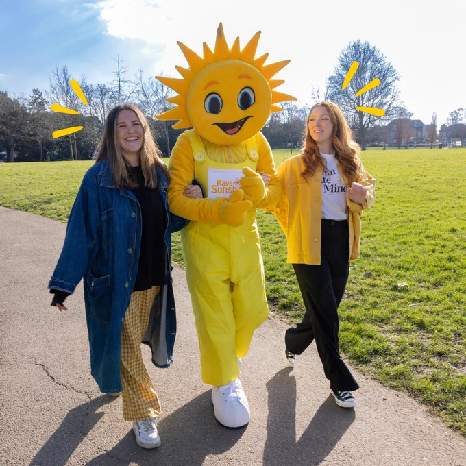 Two women walk arm in arm with a bright yellow sunshine-head madcot. The woman on the left wears yellow trousers and a long denim coat. The woman on the right wears a yellow jacket and black jeans. It is a very sunny day. 