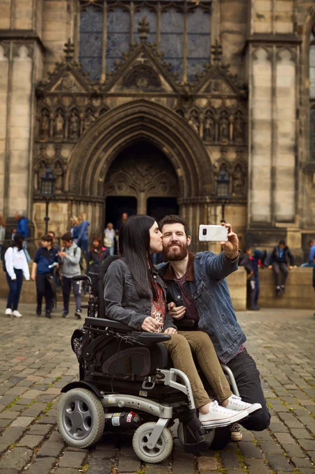 Background of image is of the ornate stone entrance to the historic St Giles Cathedral in Edinburgh. At the forefront of the image is a white female with long dark straight hair sitting in a powerchair. Kneeling next to her is a white male with dark hair and a dark beard. He is taking a selfie with his mobile phone as she kisses him on the cheek.