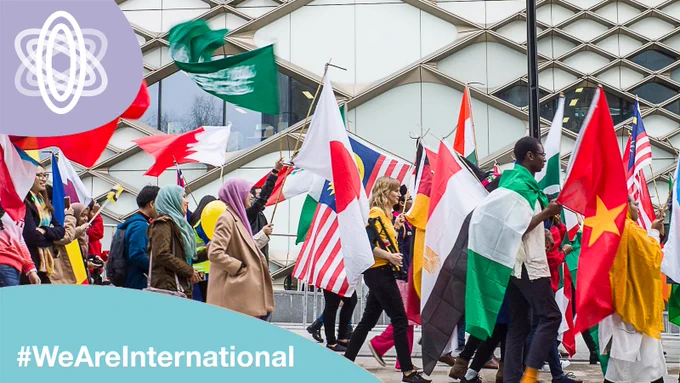 Students carrying flags walking past the Diamond building.  Text '#WeAreInternational' in one corner, We Are International logo in another corner