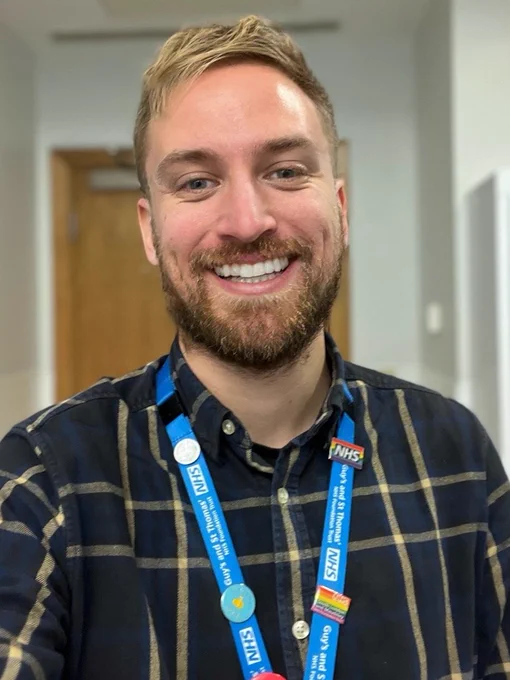 Darren is wearing a black shirt with brown vertical and horizontal stripes. He is also wearing a sky blue coloured NHS lanyard with various badges attached.