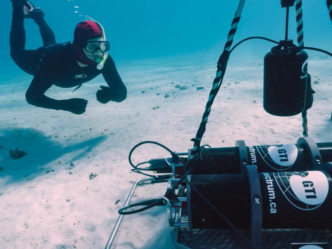 A freediver is on a sandy ocean bottom looking at an underwater microphone setup