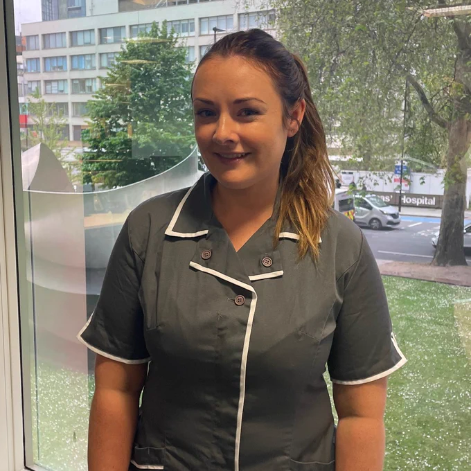 Rachael is wearing a grey short sleeved uniform with white trim around the collars, sleeves and front. Behind her is St Thomas' Hospital and vehicles are driving along Lambeth Palace Road.