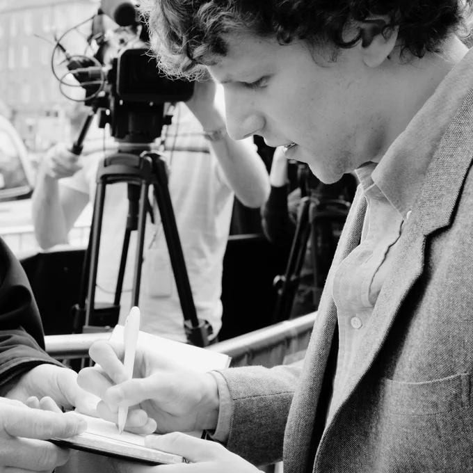 Actor Jesse Eisenberg, wearing a suit, giving interviews and signing autographs on the red carpet for Adventureland at the Cineworld. There is a film camera behind him.