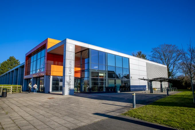 The image shows witham leisure centre, which is a building with glass and some orange bricks. Also shown are two people talking.
