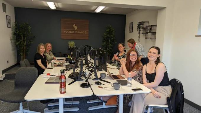 Image of 6 women sat at a long white desk covered in desktop computers smiling at the camera. In the background there is a large wall with a wooden sign with the words 'Work Space' written. The room is large, filled with lots of plants and comfortable seating.