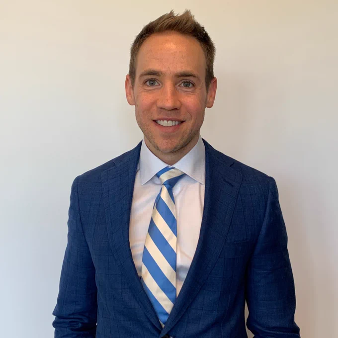 Professor Nicholas Rule stands in front of a white wall, wearing a blue suit, white dress shirt, and a blue-striped tie. 