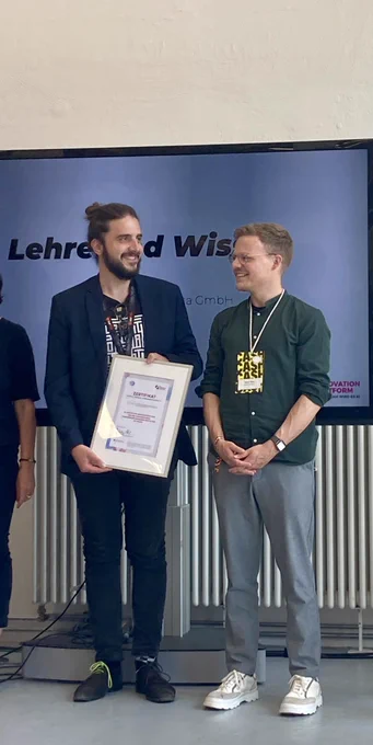 Das Bild zeigt Tobias und Moritz vor einem Fernsehbildschirm. Tobias hält ein eingerahmtes Zertifikat in den Händen.