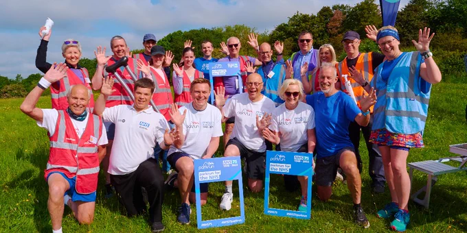 Ruth May and Steve Powis are crouched amongst a group of 18 people in a park. The group shows organisers, volunteers and runners wearing NHS branded clothing and high visibility vests. 