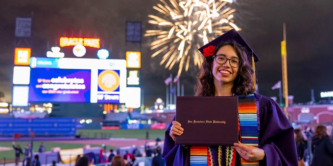 A graduate poses with their diploma cover in the stands at Oracle Park while fireworks explode behind the scoreboard